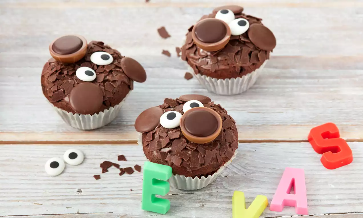 Schoko-Bärenmuffins mit runden Ohren und Augen aus Schokolade und Zucker, arrangiert auf hellem Holz mit bunten Buchstaben und Zuckeraugen