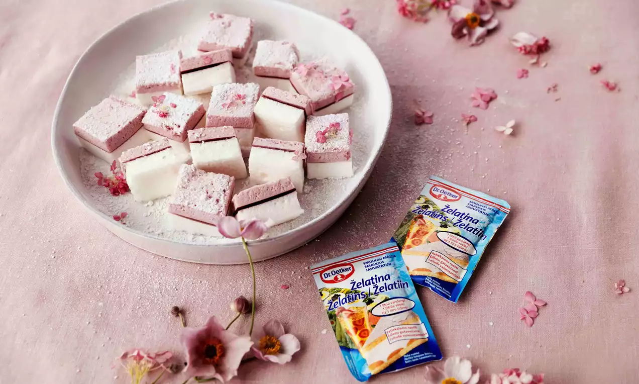 Kvadrātveida, maigi rozā un baltas krāsas zefīra gabaliņi uz balta šķīvja, pārkaisīti ar pūdercukuru, blakus gaiši rozā ziediem uz auduma fona