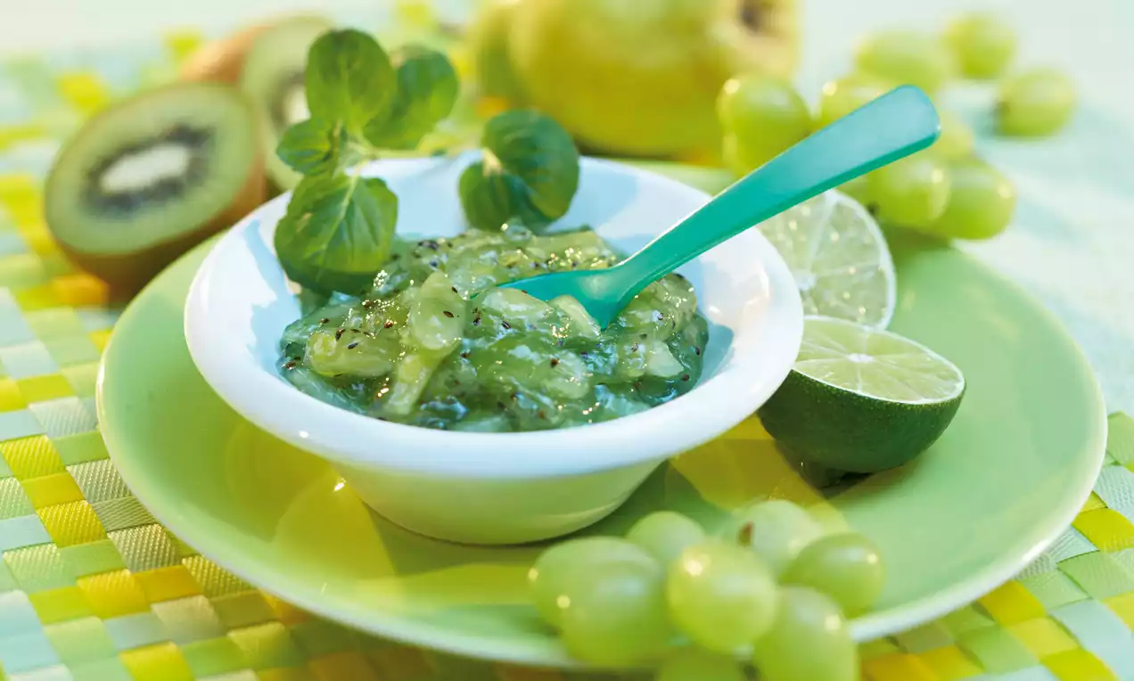 Confettura verde brillante con pezzi di kiwi e pera, servita in una ciotola bianca su un piatto verde, decorata con lime e uva