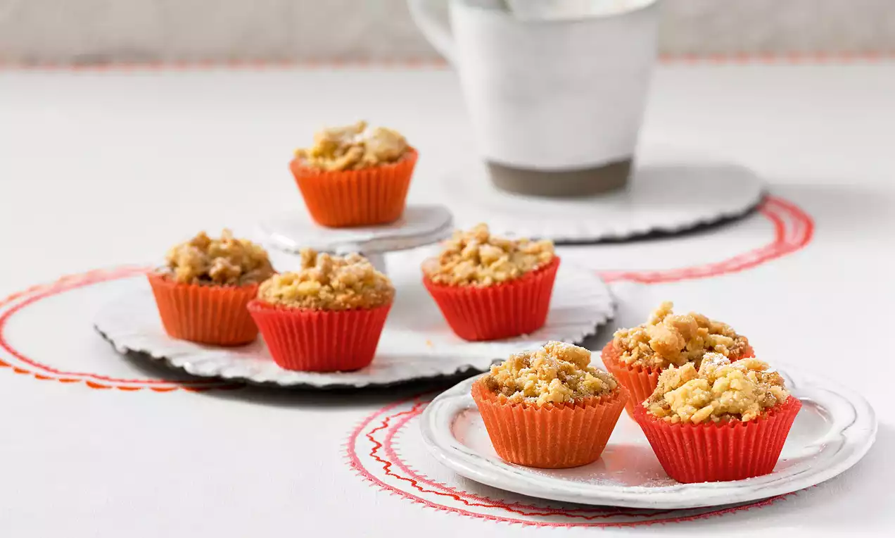 Mini-Streusel-Muffins in roten Förmchen, goldbraun und krümelig, auf weißen Tellern mit zartem Muster arrangiert, im Hintergrund ein Becher