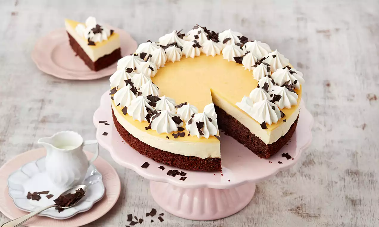 Troslojna torta s tamnobrownom biskvitnom podlogom, svijetlim kremastim punjenjem i žutom glazurom, obrubljena hrpicama šlaga i čokoladnim strugotinama na pastelno roze tanjiru