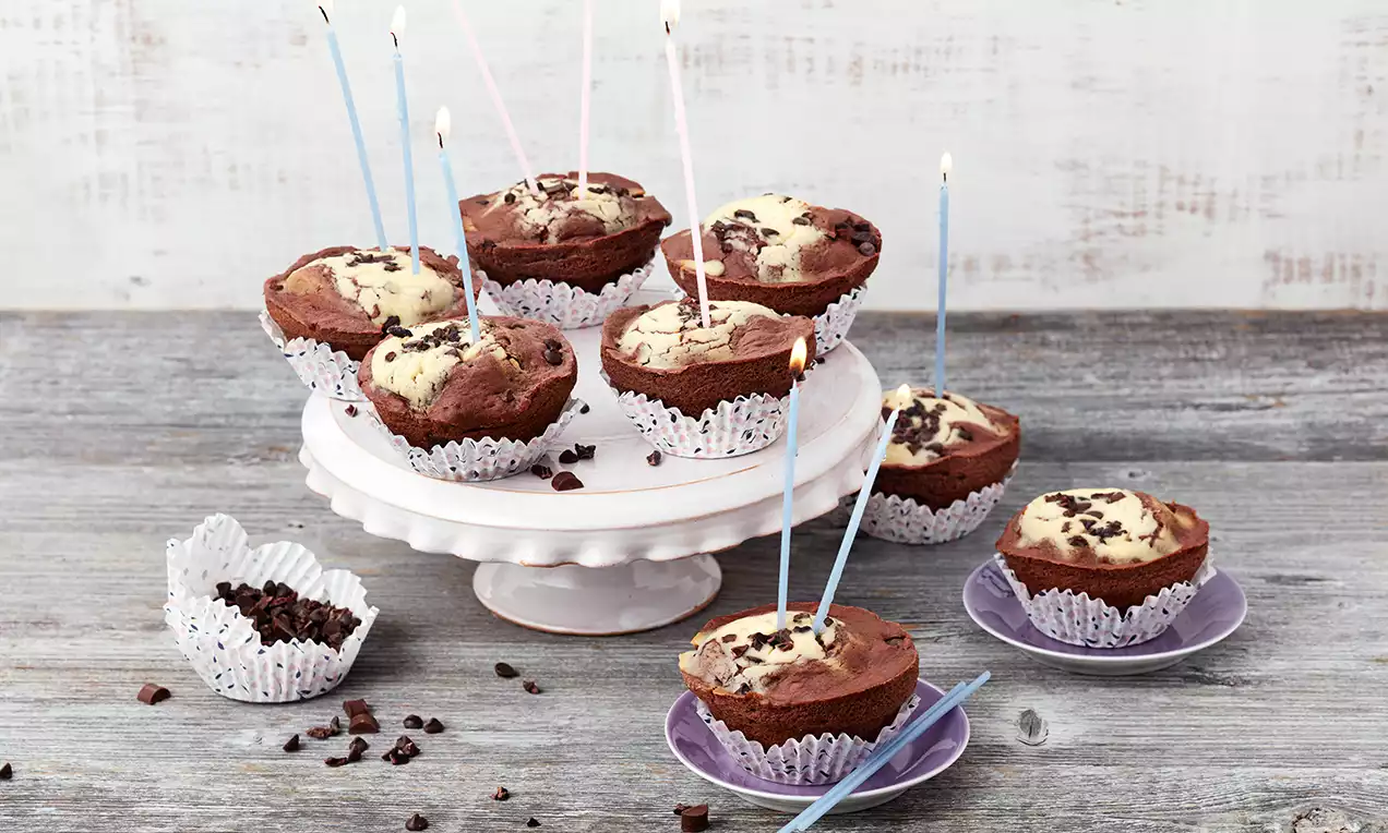 Schoko-Cheesecake-Muffins mit zweifarbiger Marmorierung, dekoriert mit Schokostückchen und Kerzen, serviert auf einer weißen Tortenplatte