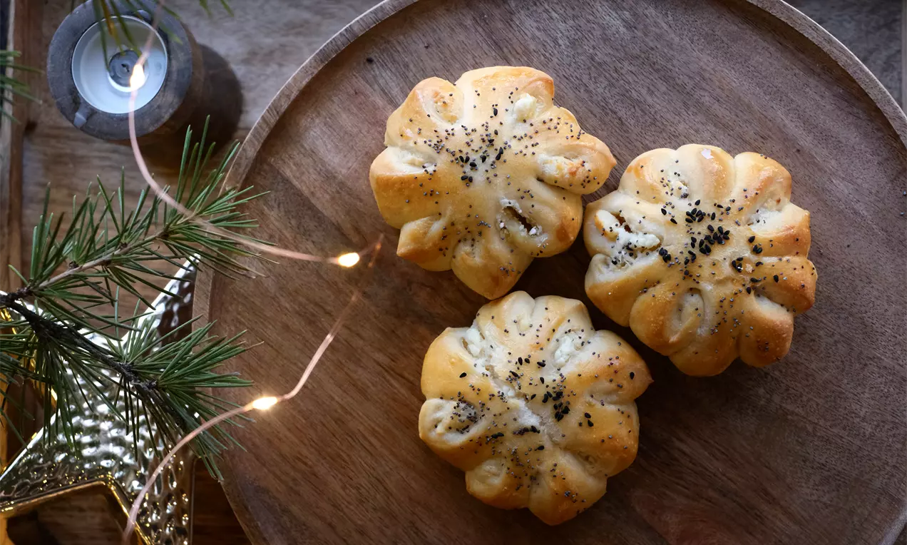Златистокафяви хлебчета с форма на разцъфнали цветя, поръсени със семена мак на дървена подложка до елхови клонки и светеща декорация