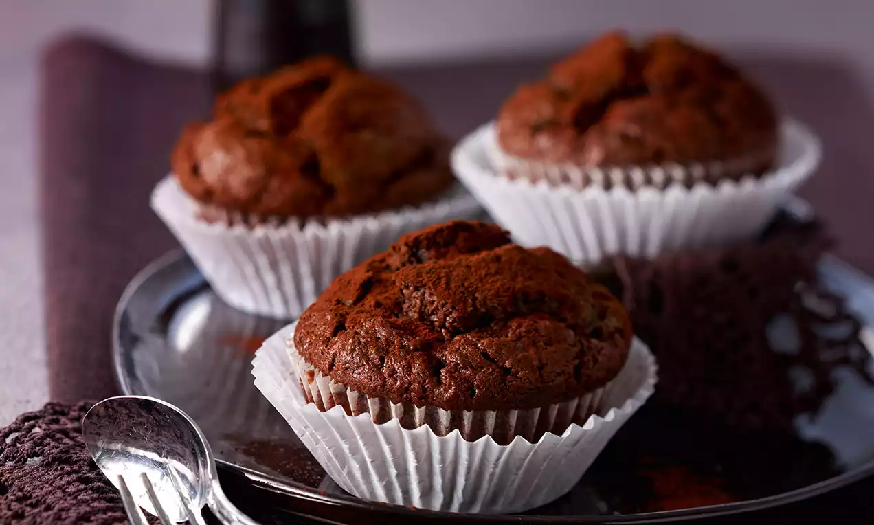 Dunkelbraune, lockere Schokoladen-Zucchini-Muffins in weißen Papierförmchen auf einem glänzenden Teller mit lilafarbenem Stoff drapiert