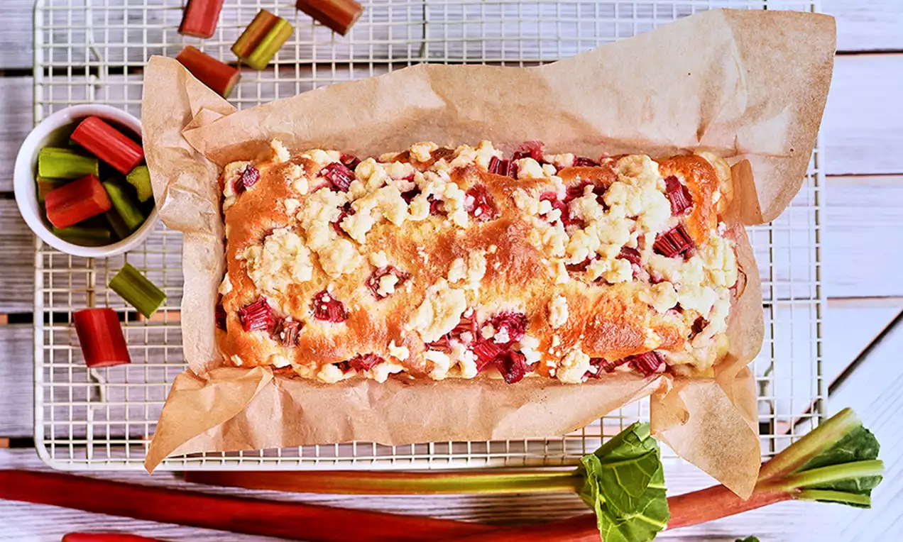 Złocista podłużna babka drożdżowa w papierze, z soczystymi kawałkami rabarbaru i maślaną kruszonką, na kratce obok łodyg rabarbaru