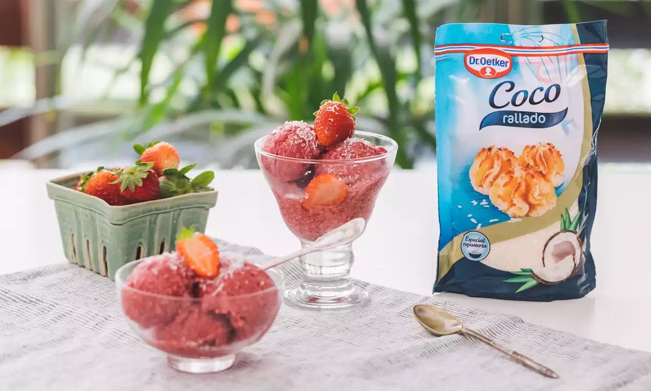 Helado rosado y cremoso de fresas, coco y plátano en copas de cristal, decorado con fresas frescas sobre un mantel claro