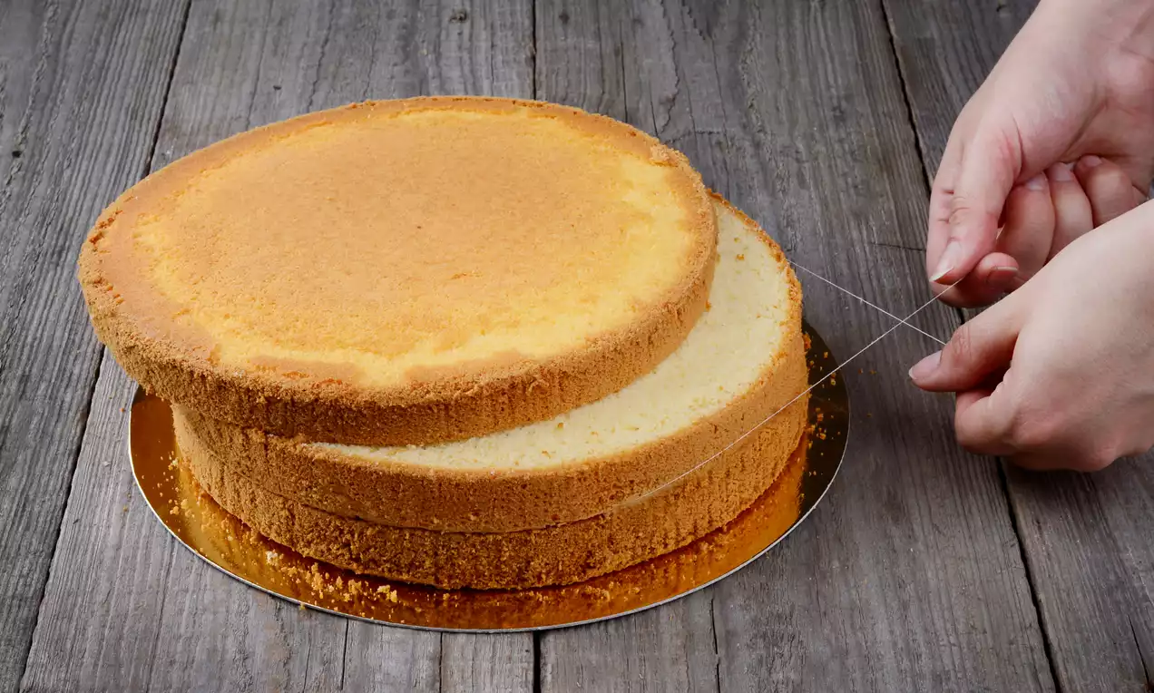 Jasnozłoty biszkopt o puszystej fakturze, przecinany nitką na równe warstwy, na złotej podkładce, na tle surowego drewnianego blatu