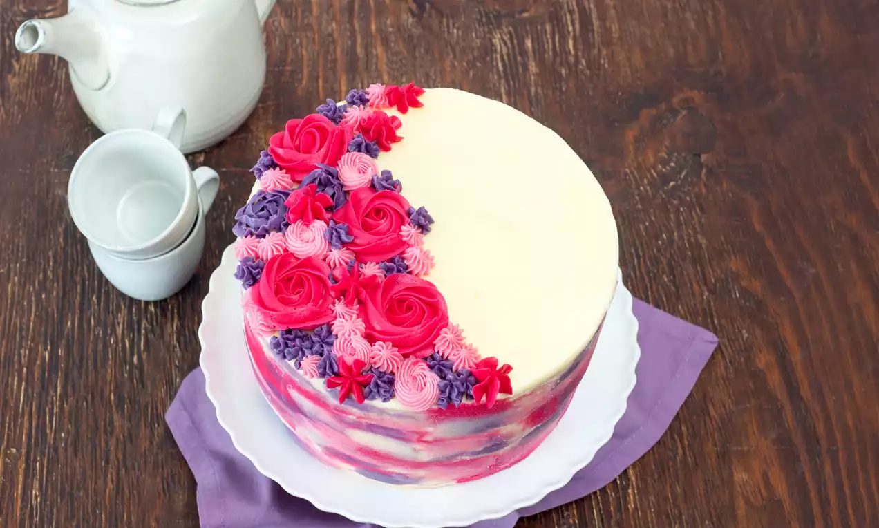 Runder, pastellfarbener Kuchen mit cremeweißer Oberfläche und einer Seite aus rosa und lila Blumen auf Teller mit lila Stoff drapiert