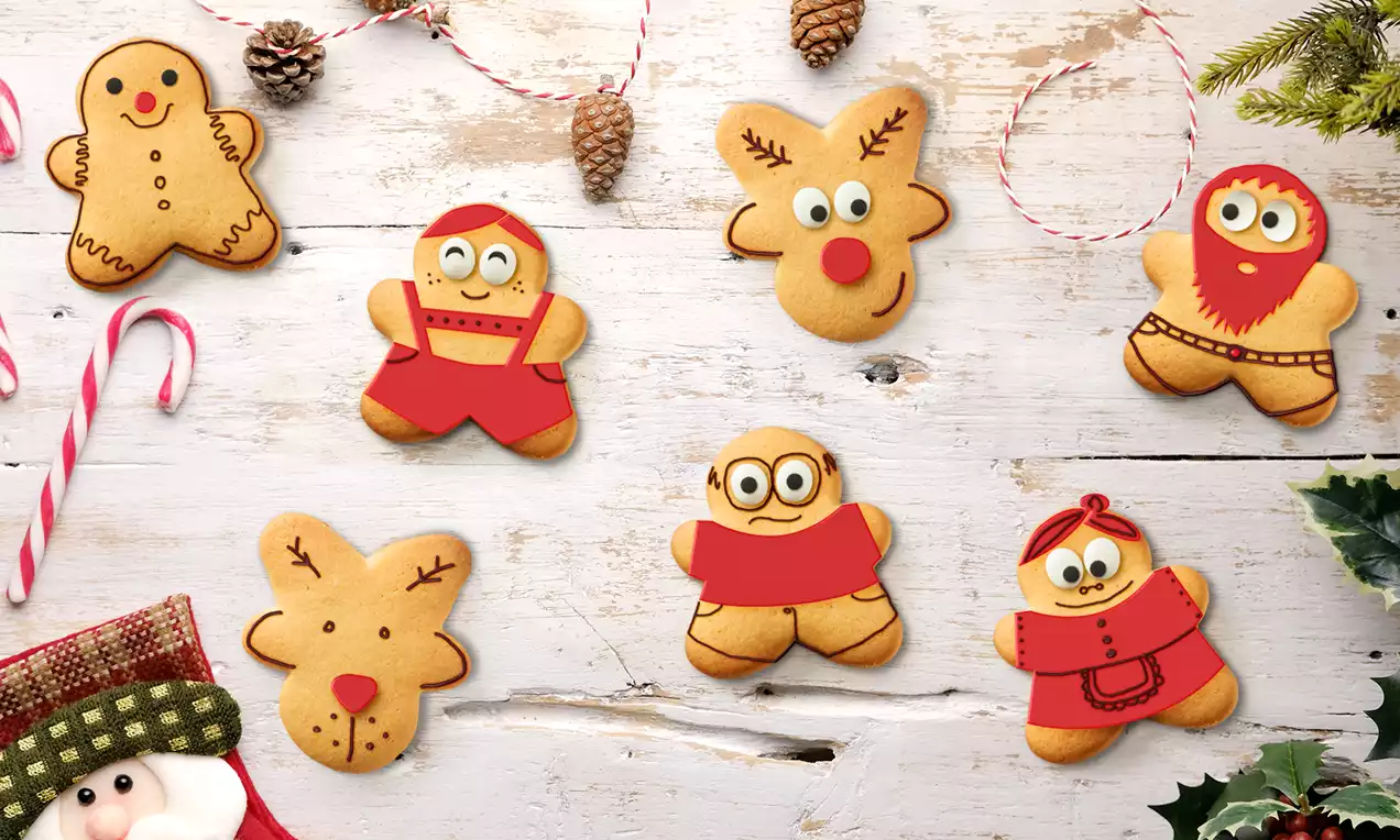 Galletas doradas con forma de personajes navideños sobre mesa blanca de madera, rodeadas de bastones, piñas y ramas verdes