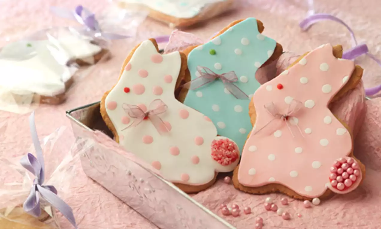 Galletas con forma de conejito con glaseado rosa, azul y blanco decoradas con puntos, lazos y perlitas en una bandeja metálica