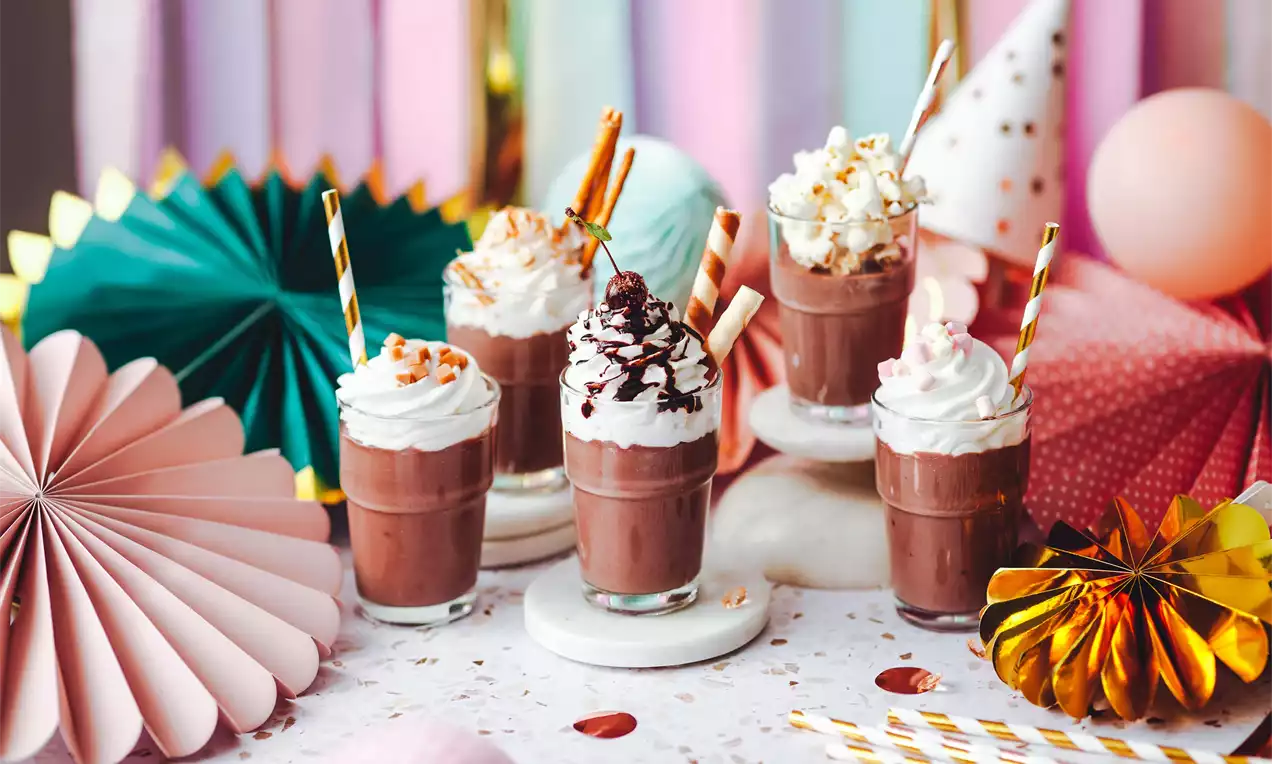 Pět sklenic čokoládového pudinkového drinku zdobených šlehačkou, křupkami, čokoládovou polevou a slánou dekorací na barevném stolním aranžmá