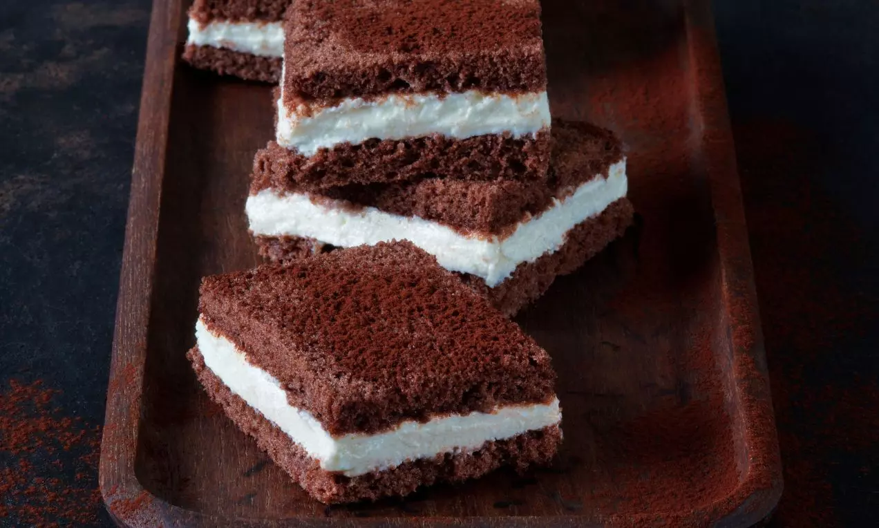 Tmavě hnědé obdélníky z nadýchaného kakaového těsta s jemnou bílou vrstvou krému uvnitř, poskládané na dřevěné podnose