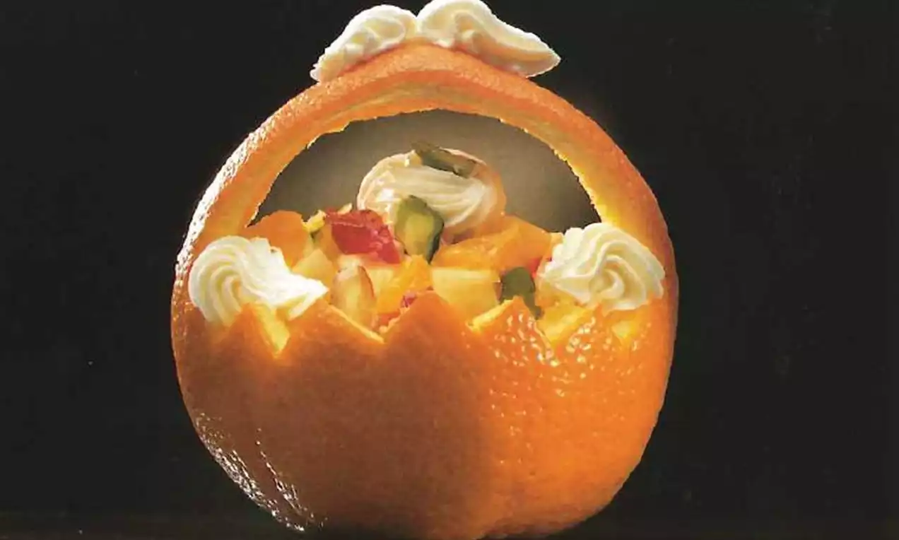 Panier sculpté dans une écorce d’orange brillante, rempli de fruits colorés et décoré de rosettes de crème blanche