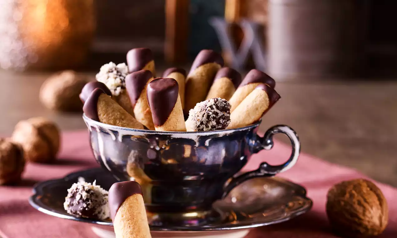Walnuss-Makronen-Stäbchen mit dunkler Schokoladenspitze in einer glänzenden Tasse auf rosa Stoff, umgeben von Walnüssen und Trüffeln