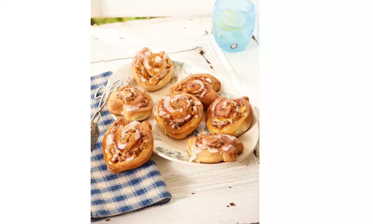 Hrskavi zlatnosmeđi pužići sa punjenjem od jabuka i preliveni bijelom glazurom, posluženi na bijelom tanjiru uz plavo karirano platno