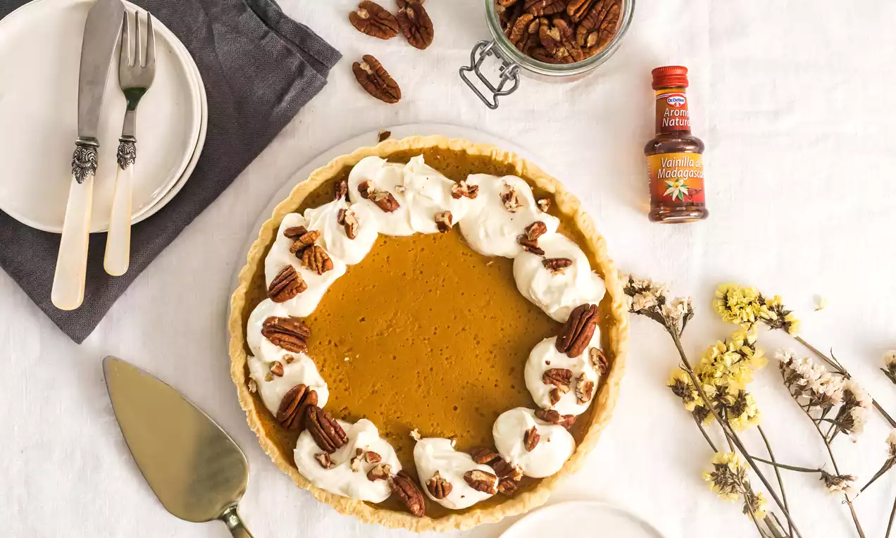 Tarta circular de calabaza color naranja intenso con bordes crujientes, decorada con montañas de nata blanca y mitades de nuez sobre un mantel claro