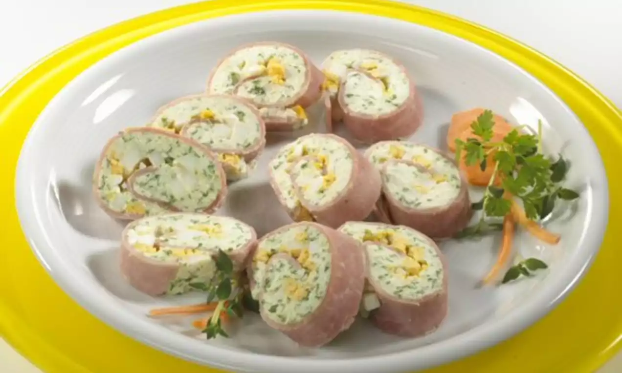 Rosa Schinkenröllchen mit weiß-grüner Kräuterfüllung und gelbem Ei auf ovalem Teller, dekoriert mit gelber Möhre und Petersilie