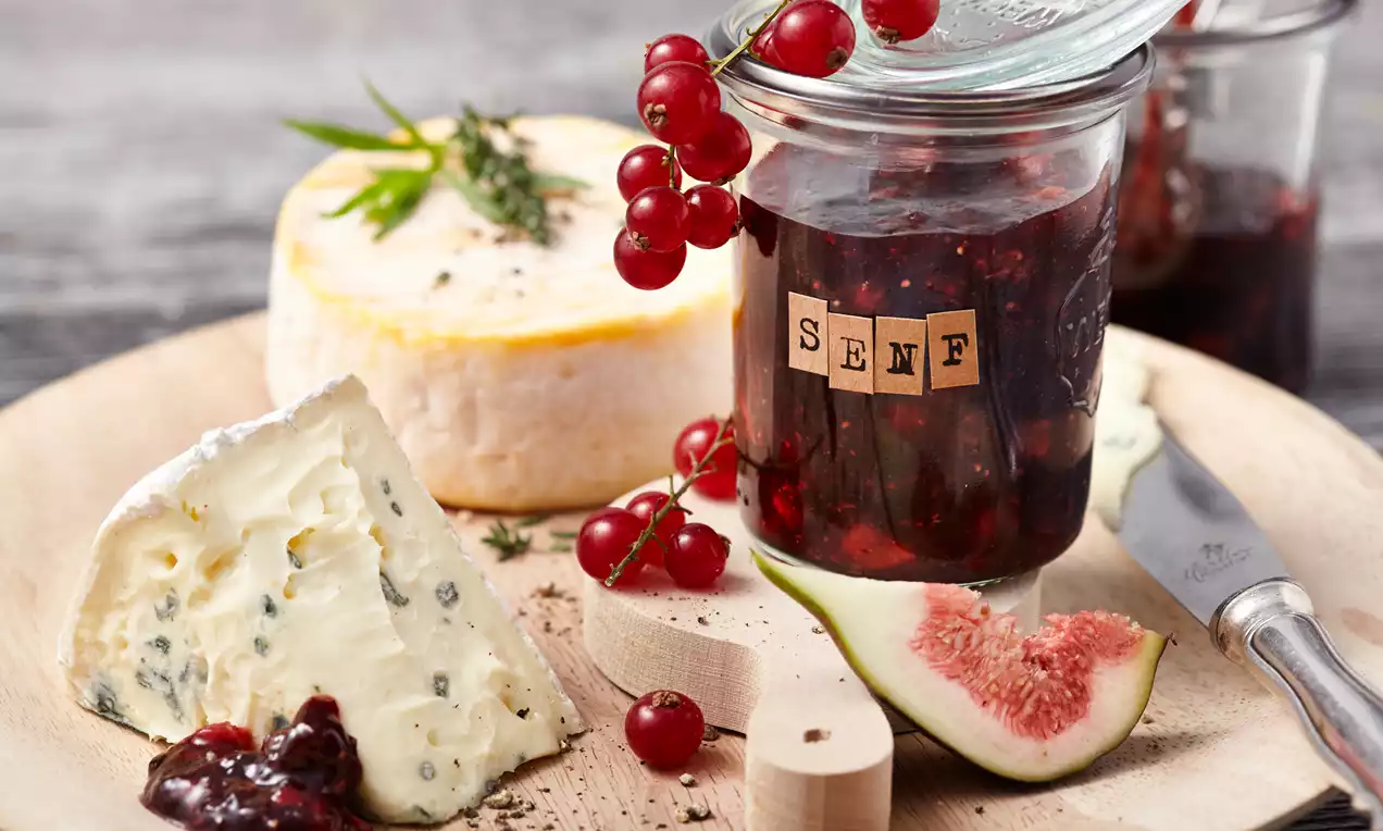 Ein Glas mit tiefrotem Feigen-Johannisbeer-Senf, garniert mit roten Johannisbeeren, daneben Käse, frische Feige und ein Holzlöffel auf einem Holzbrett