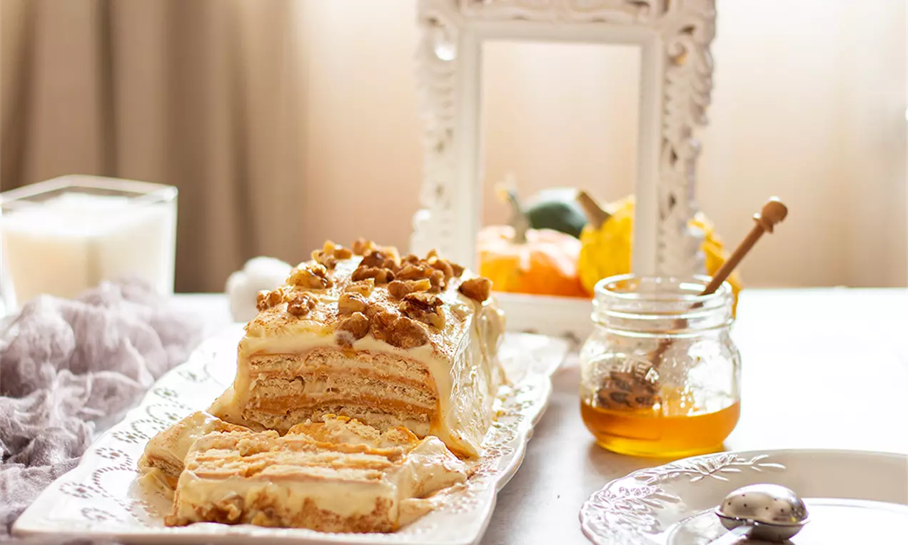 Бисквитена торта с пластове светложълт ванилов крем и тиква, покрита с натрошени бисквити, сервирана върху правоъгълна чиния
