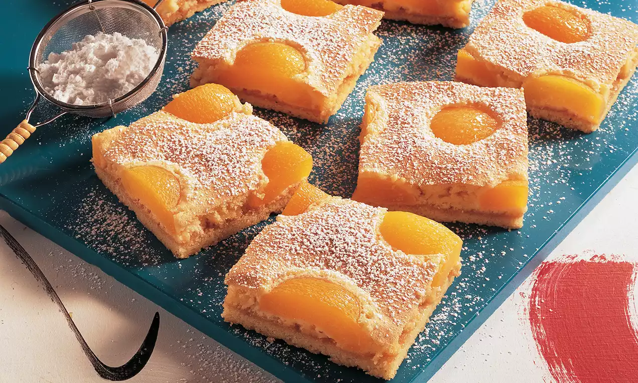 Goldgelber Marillenkuchen in rechteckigen Stücken mit halbierten, glänzenden Aprikosen und Puderzucker bestäubt auf dunkelblauem Teller