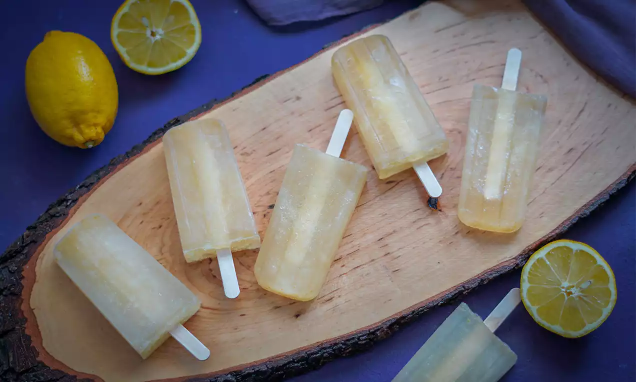 Hellgelbe Zitronen-Daiquiri-Eisstiele mit transparenter, leicht glänzender Oberfläche, angeordnet auf hellem Holzbrett mit frischen Zitronen
