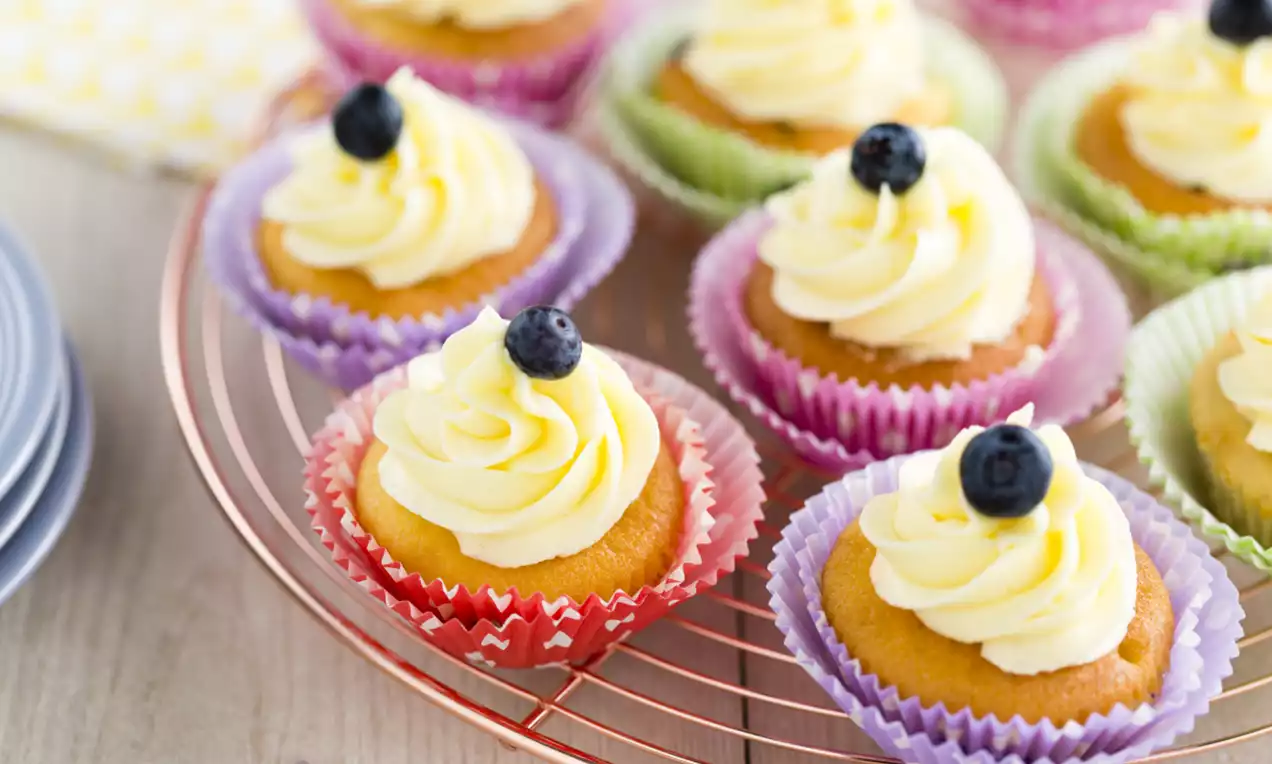 Goldene Cupcakes mit cremigem, weißem Yoghurttopping und einzelnen dunkelblauen Blaubeeren in bunten Papierförmchen auf einem Roségold-Gitter