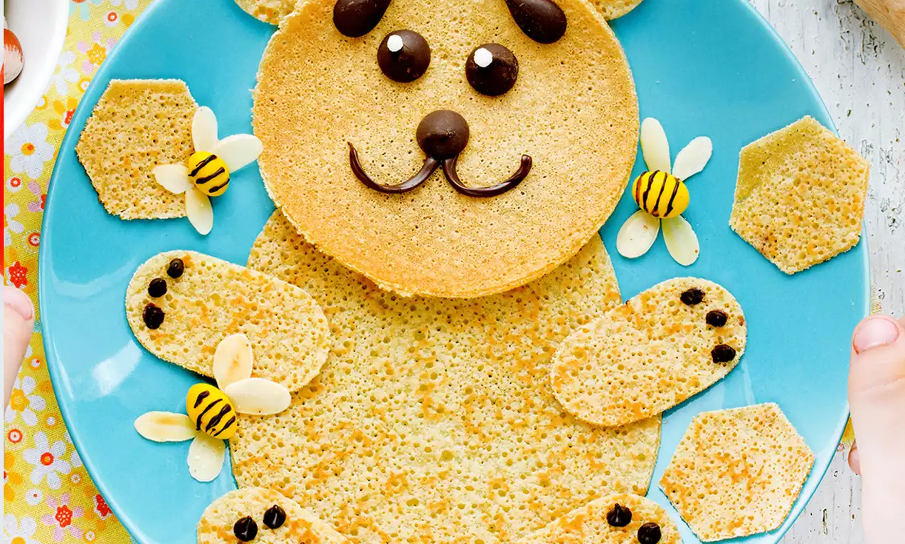 Crêpes dorées en forme d'ourson avec détails en chocolat et amandes, accompagnées de motifs d'abeilles et de nids d'abeilles sur une assiette bleue
