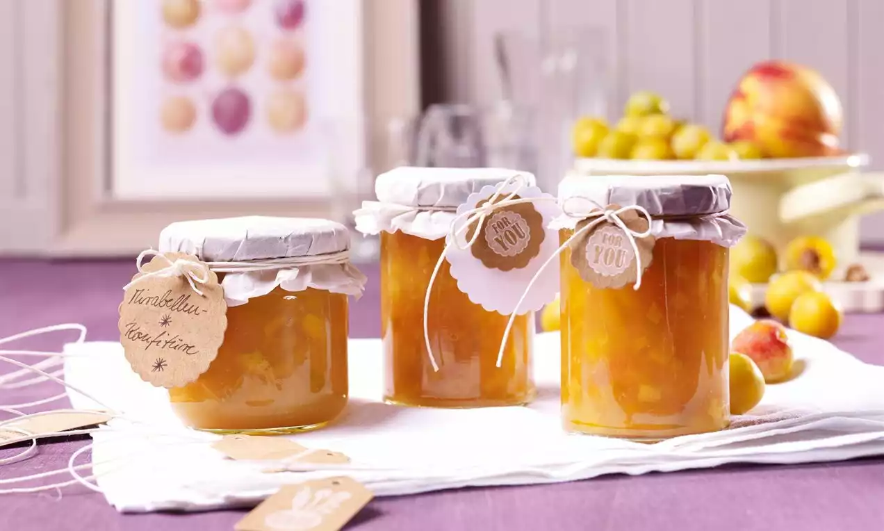 Tři sklenice mirabelkového džemu s papírovými víčky, lesklá zlatavá struktura, ozdobné cedulky, na bílém ubrusu mezi čerstvým ovocem