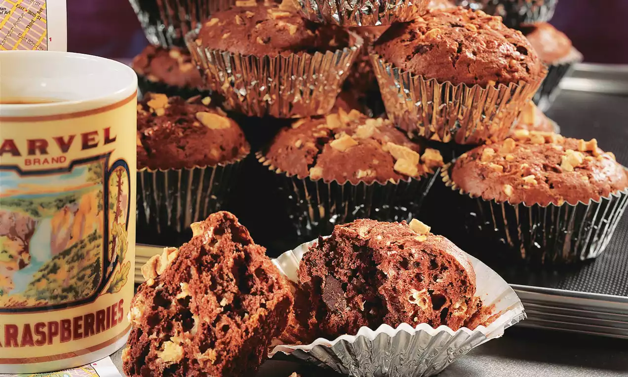 Muffin al cioccolato scuro con pezzi di mandorla dorata, texture morbida e umida, avvolti in pirottini argentati su un vassoio