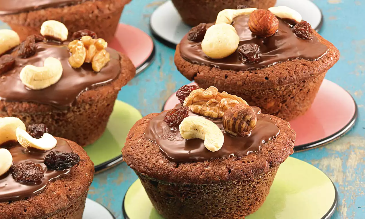 Muffin marrone cioccolato decorati con una glassa lucida e guarnizioni di noci miste e uvetta, disposti su piattini colorati