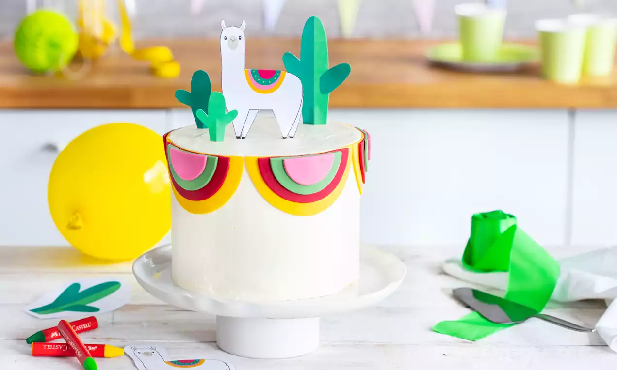 Lamas kūka ar gludu baltā krāsā pārklātu virsmu, rotāta ar krāsainiem pusapļu motīviem un papīra dekorācijām – lamu un zaļiem kaktusiem