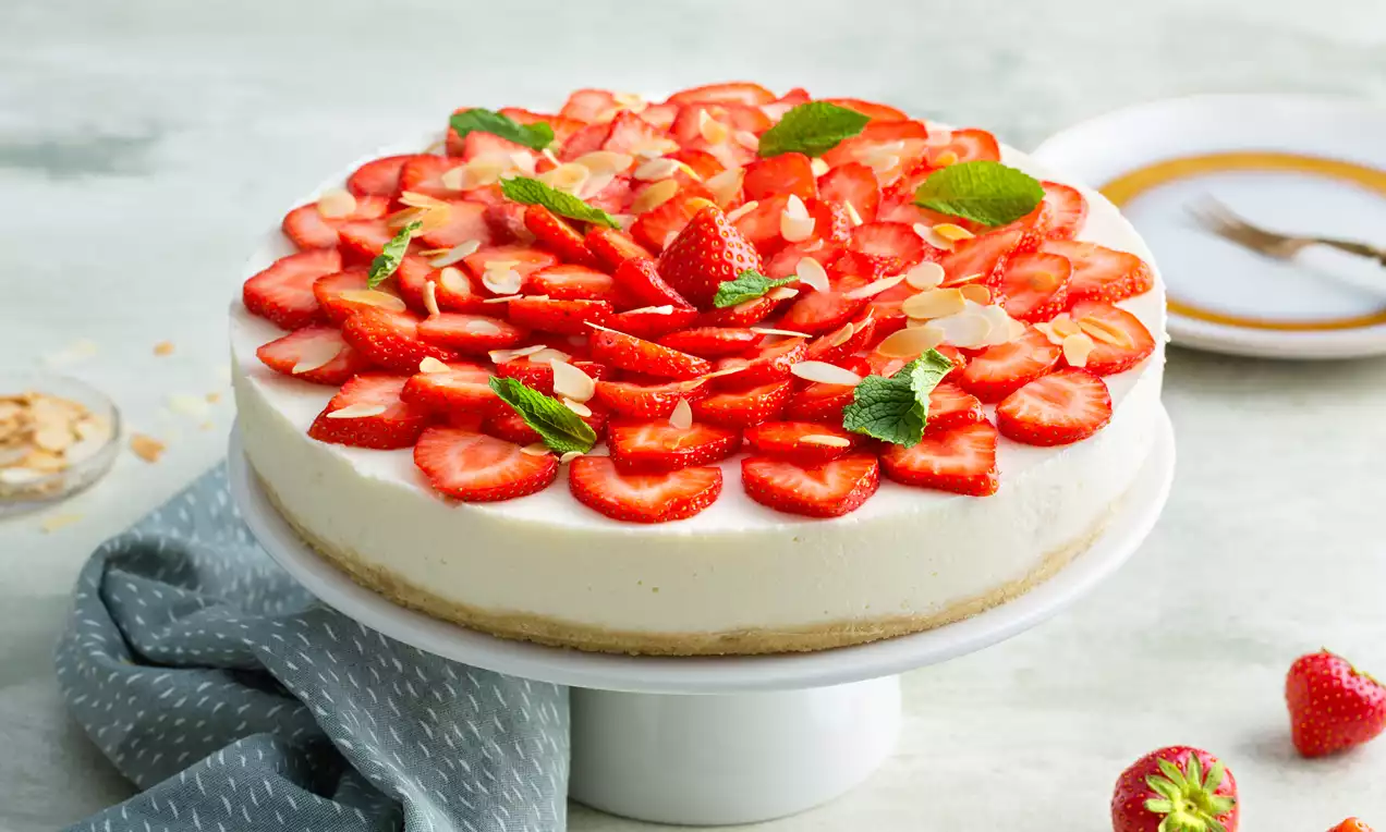 Kwarktaart met een lichte, gladde vulling op een zandkleurige bodem, versierd met rode aardbeien, groene munt en amandelschaafsel op een wit taartplateau