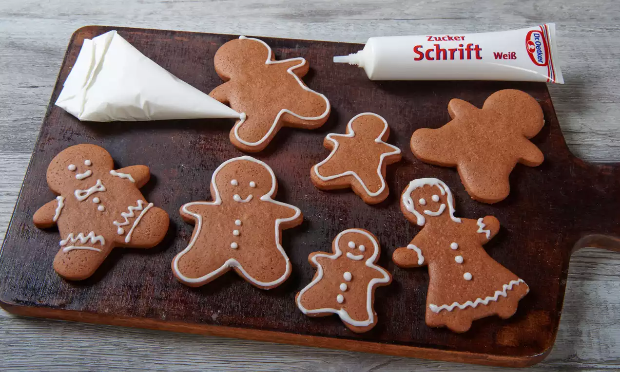 Mehrere braune Lebkuchenfiguren mit weißem Zuckerguss dekoriert auf einem dunklen Holzbrett neben Spritztüte und Zuckerschrift-Tube