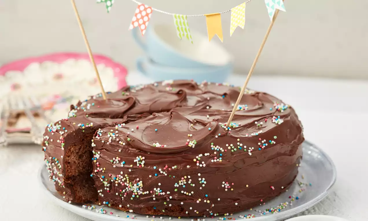 Rundlicher Schokoladenkuchen mit glänzender Glasur, bunten Streuseln und bunten Wimpeln auf einem Teller mit hellen Hintergrundelementen