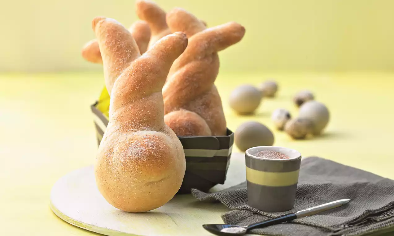 Goldbraune Dinkel-Häschen mit weicher Oberfläche und gebogenen Ohren, arrangiert in einem Streifenkorb auf hellem Untergrund, daneben Eier und Espresso