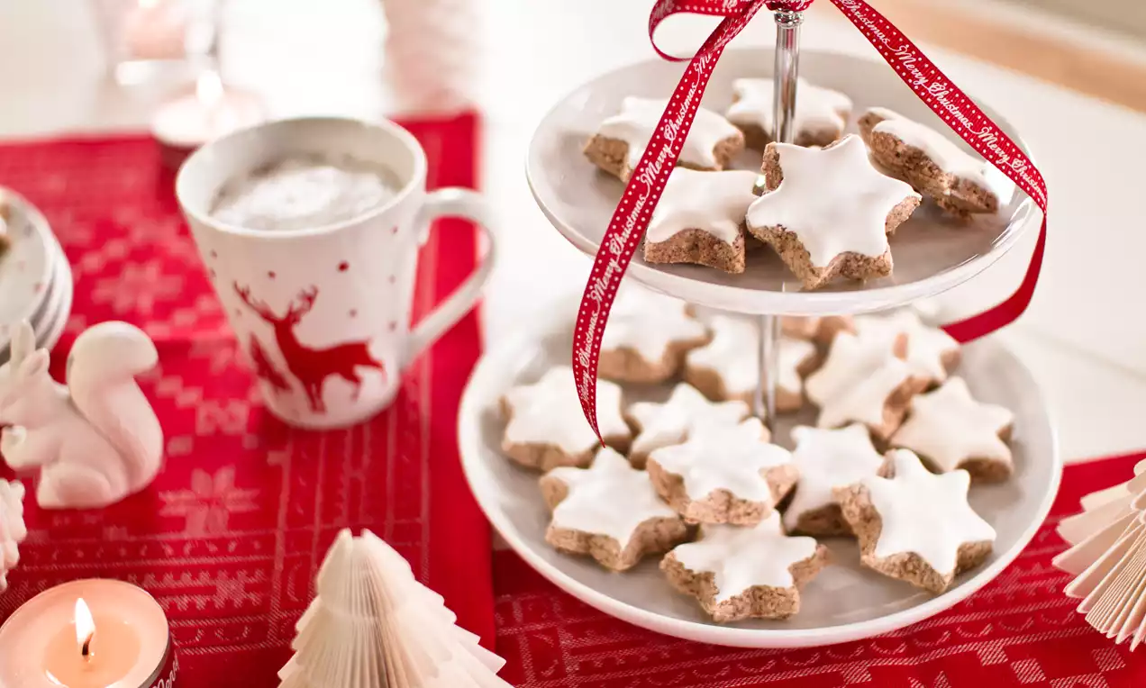 Zvjezdasti kolačići s bijelom glazurom na dvokatnom staklenom tanjuru s crvenom satenskom vrpcom, okružen božićnim ukrasima na crvenom stolnjaku