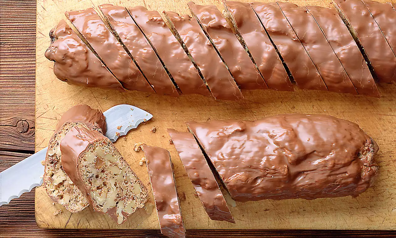 In Scheiben geschnittener, mit glänzendem Schokoladenüberzug bedeckter Nusszwieback auf einem Holzbrett mit gezacktem Messer daneben