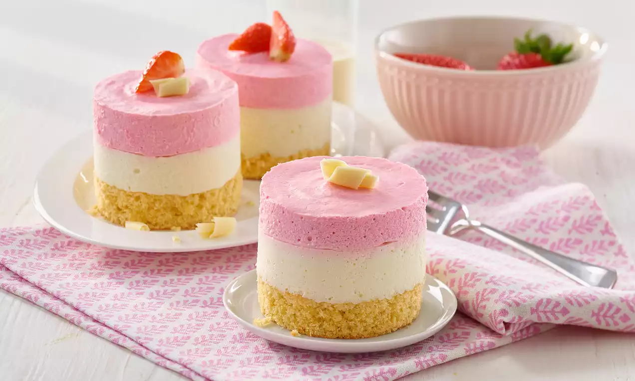 Schicht-Törtchen mit rosa Erdbeercreme, weißer Vanillecreme und hellem Keksboden, garniert mit Erdbeerstück und weißen Schokoladensplittern