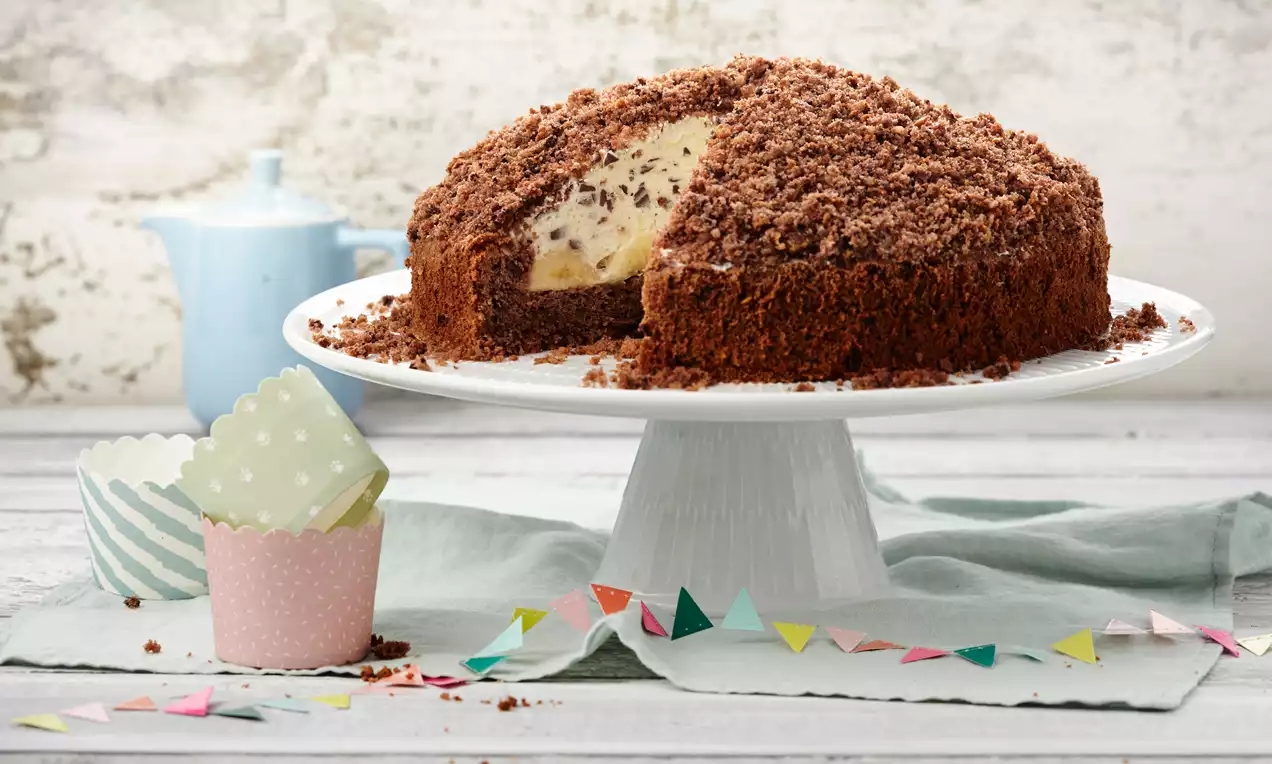 Schnittfester runder Schokoladenkuchen mit heller Sahne-Bananenfüllung, bedeckt von krümeliger Schokoschicht auf weißer Tortenplatte