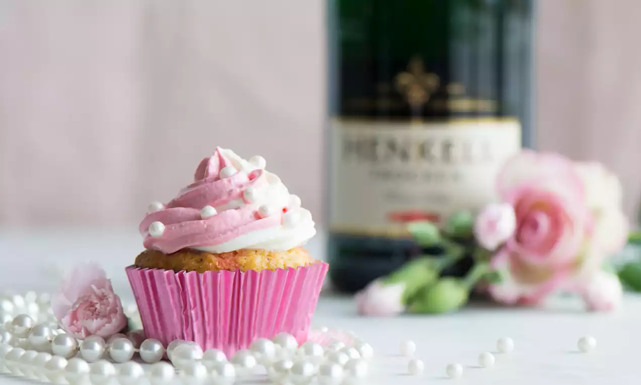 Cupcake mit rosa-weißer Cremehaube, verziert mit Perlen, in pinker Papierform, dekoriert mit Perlenschmuck und rosa Blüten vor einer Sektflasche