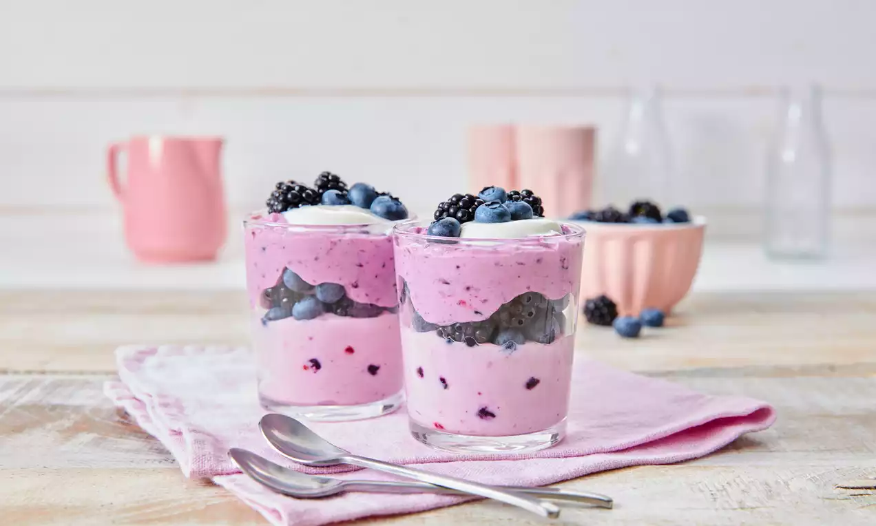 Zwei Gläser mit rosa-cremigem Waldbeeren-Dessert, geschichtet mit Beeren, auf rosa Stofftuch mit zwei Löffeln dekoriert in heller Umgebung