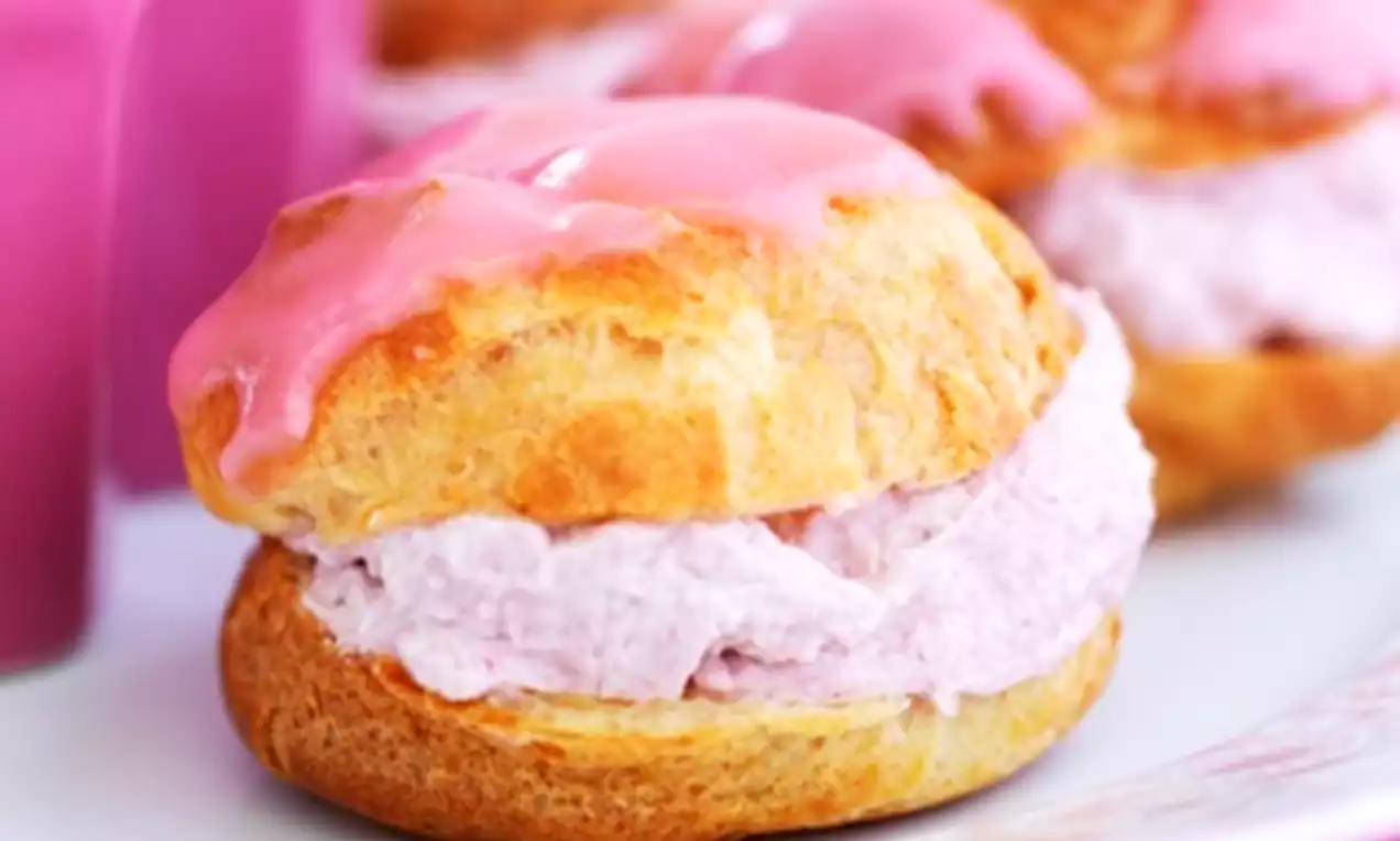 Ronde goudbruine soezen gevuld met luchtige lichtroze frambozenroom en geglazuurd met glanzend roze frosting op een wit bord