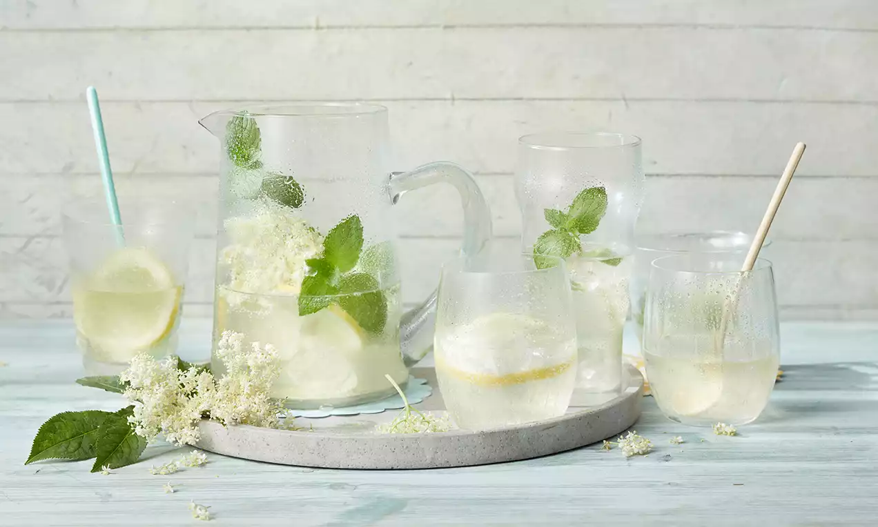 Kühle Holunderblüten-Limonade in Gläsern und einer Karaffe, mit Zitronenscheiben, Minzblättern und Holunderblüten auf hellem Tablett arrangiert