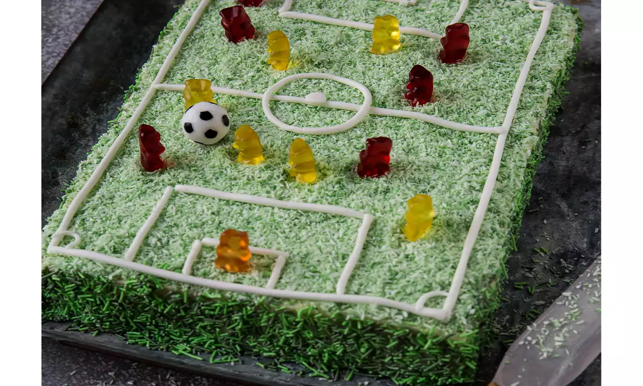 Tourte rectangulaire recouverte de copeaux de noix de coco teints en vert avec des lignes en crème blanche et des bonbons en forme de joueurs