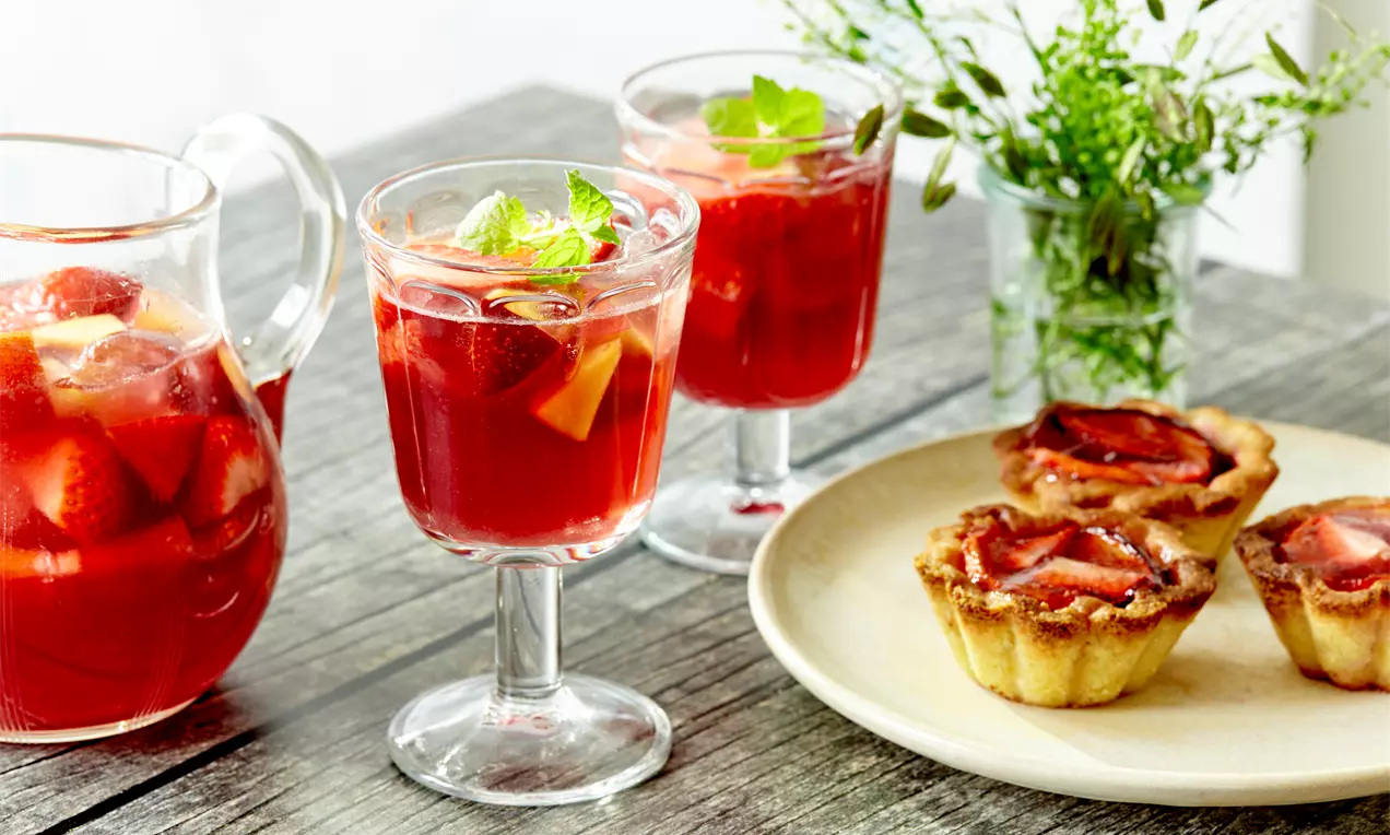 Червени напитки с парченца ягоди и цитрусови плодове в стъклени чаши, гарнирани с листа мента до кана и десерти върху светла чиния