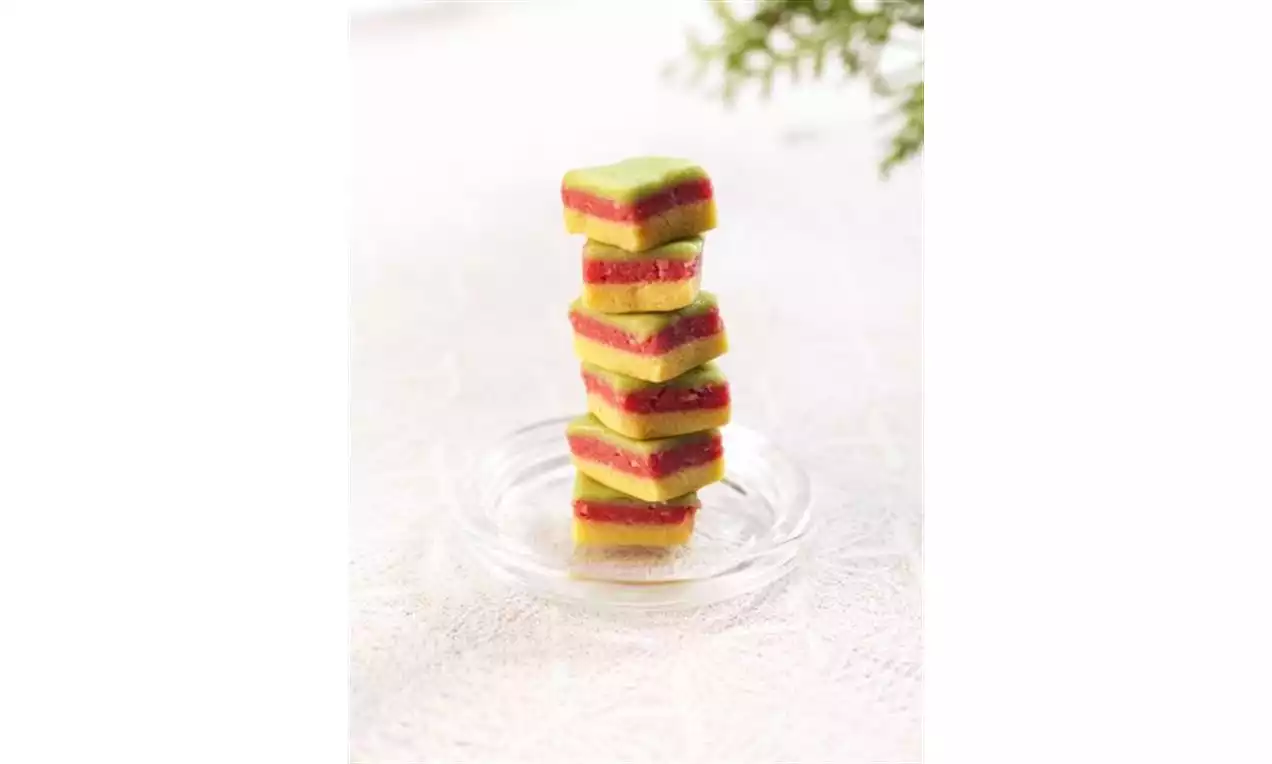 Firkantet teaterkonfekt i lag av grønn marsipan og rød fyll stablet høyt på et lite glassfat med hvit teksturert bakgrunn