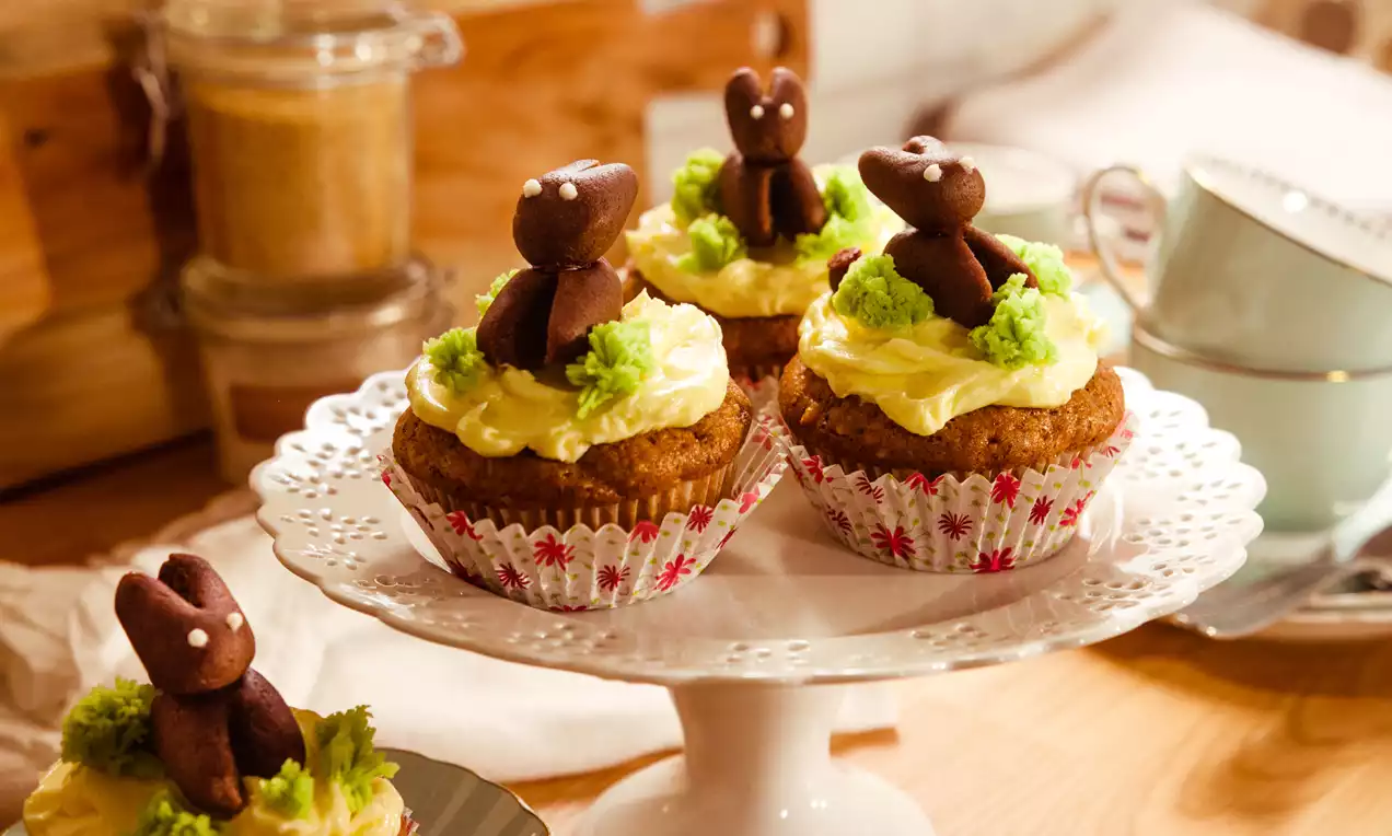 Veganer Karottenmuffin mit hellgelbem Frosting, grünen Deko-Tupfen und dunklem Schoko-Häschen auf verspielter weißer Tortenplatte