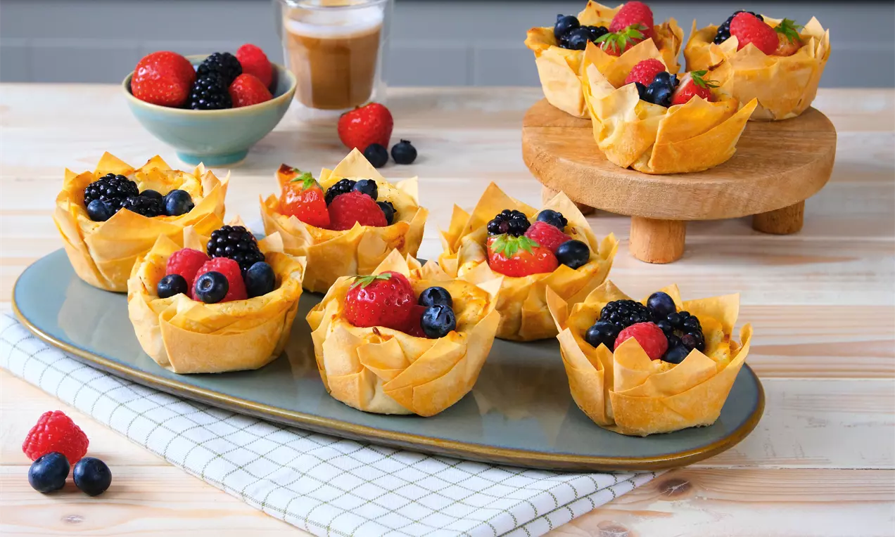 Kleine Filoteig-Körbchen mit cremiger Käsekuchenfüllung, goldbraun gebacken, bestäubt mit Puderzucker und garniert mit Himbeeren und Minze