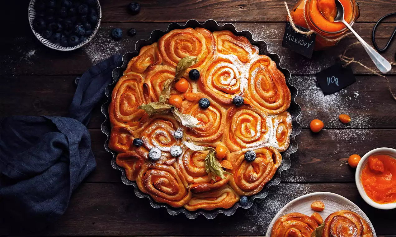 Vosí hnízda zlatohnědá a lesklá, tvořící spirálové vzory v kulaté formě, zdobená mrkvovým džemem, physalis a borůvkami na dřevěném stole