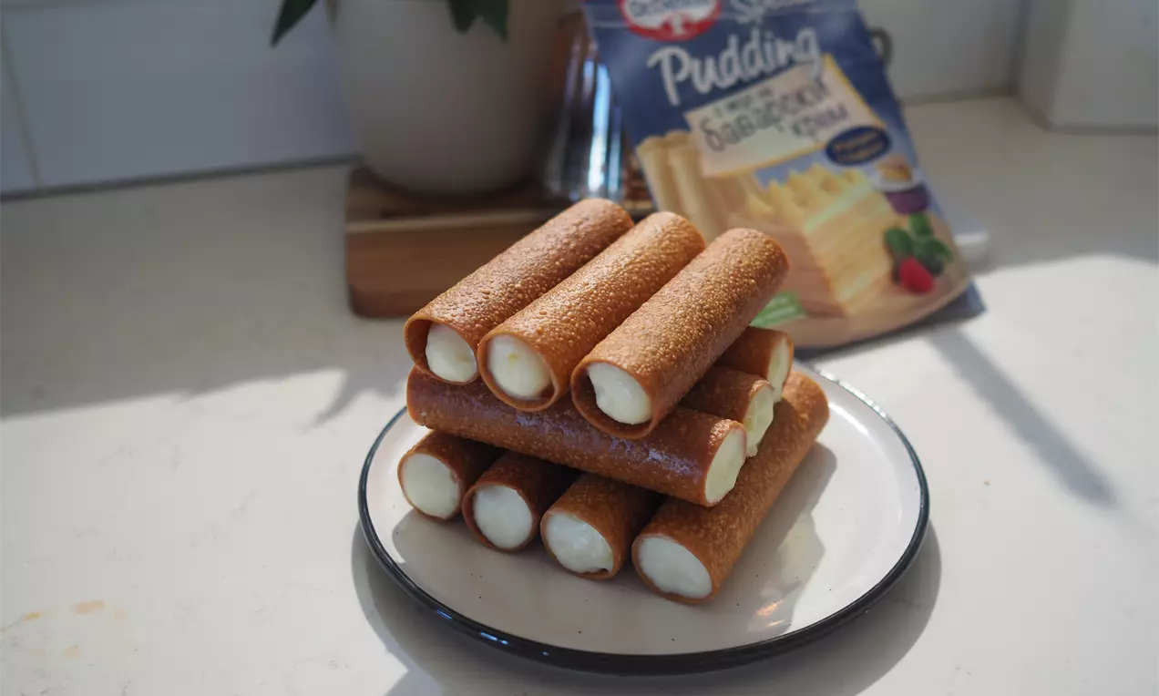 Златисто-кафяви хрупкави каноли, подредени като пирамида върху бяла чиния, пълни с гладък баварски крем, леко блестящ на светлината